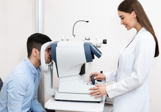 Eye Inspection Concept. Side view of smiling female optometrist ophthalmologist examining eyesight and vision of young male customer in modern clinic. Optician using apparatus for diagnosis