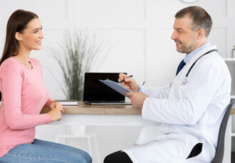 Medicine, Healthcare and People Concept. Smiling doctor with clipboard and young woman patient meeting at hospital. Specialist holding chart and writing anamnesis and symptoms