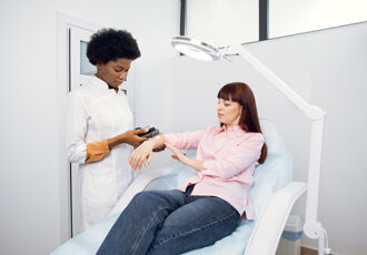Healthcare, oncology, skin cancer detection. Pleasant young black woman, doctor dermatologist looking at female patient's mole on hand with modern digital magnifier dermatoscope